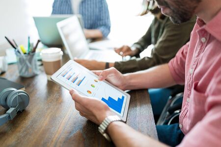 Businessman examining graphs on digital tablet while planning with coworkers at desk in officeの写真素材