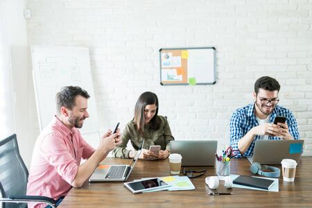 Happy business people using internet on smartphone while ignoring work at officeの写真素材