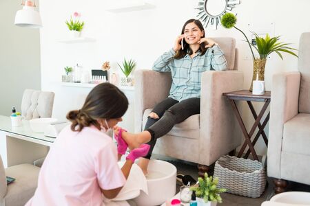 Smiling young customer talking on smartphone while beautician massaging her foot at salonの写真素材