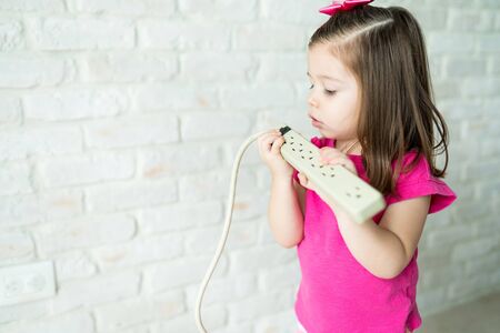Curious cute female toddler looking at electric cable while standing by wall in living room at home