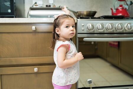 Cute female toddler standing by oven in kitchen at homeの写真素材