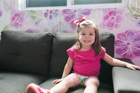 Smiling small cute girl sitting on sofa in living room at homeの写真素材
