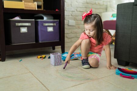 Adorable little girl drawing with colorful crayons on tiled floor at homeの写真素材