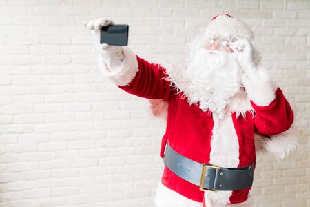 Man in Santa costume holding glasses while taking selfie through smartphone against white brick wallの写真素材