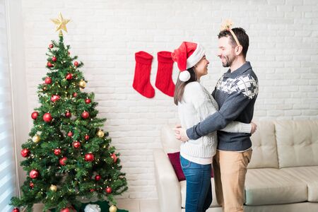 Loving happy couple embracing while standing by Christmas tree at homeの写真素材