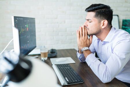 Handsome young Latin finance professional sitting with eyes closed while hoping for good stock trade...