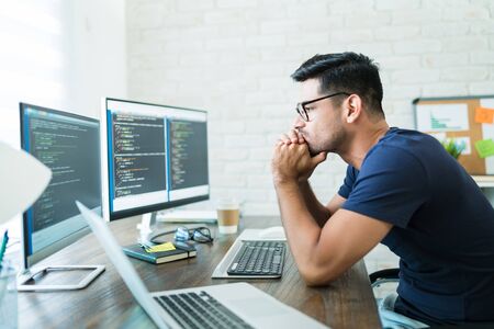 Thoughtful young handsome programmer analyzing software codes on computer screens while working from...