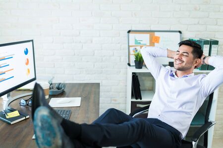 Full length of young male Latin sales freelancer relaxing with feet up on deskの写真素材