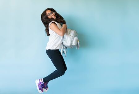 Full length of excited young girl holding backpack while jumping against plain background