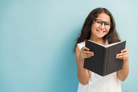 Happy Hispanic female student with eyeglasses holding book while making eye contact against...