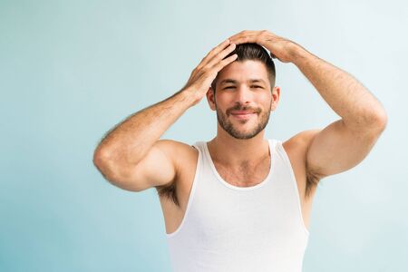 Portrait of attractive smiling muscular man with hands in hair while grooming against turquoise backgroundの写真素材