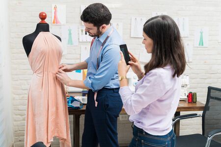Female dressmaker photographing from smartphone while male coworker dressing dummy at workshopの写真素材