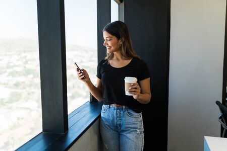 Smiling female executive using smartphone while holding coffee cup by window at office
