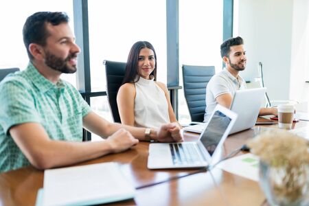 Beautiful young businesswoman making eye contact while sitting amidst colleagues at desk during...