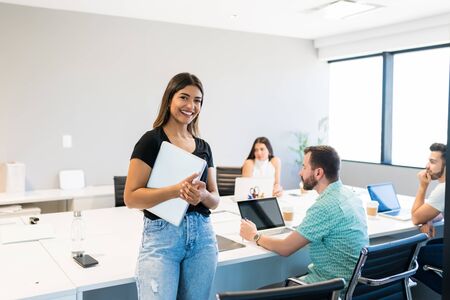 Confident smiling female professional standing with laptop at desk in workplace