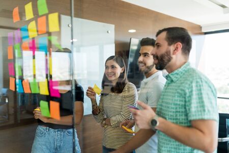 Male and female colleagues arranging adhesive notes on glass wall in meeting at officeの写真素材