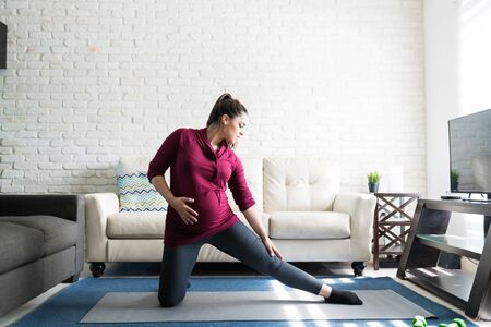 Attractive expectant woman kneeling while stretching her leg on mat against sofa at home