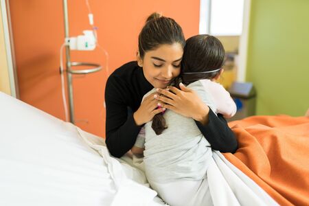 Young woman embracing unwell daughter on hospital bed during treatment