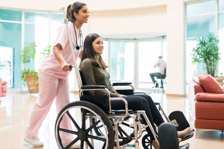 Female healthcare worker pushing recovered woman on wheelchair in hospital lobbyの写真素材