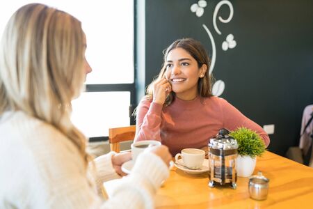 Smiling Latin woman listening to female friend while having coffee at table in restaurantの写真素材