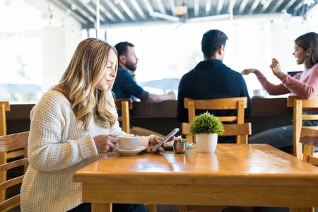 Caucasian woman using smartphone while having coffee at table with friends sitting in background at...