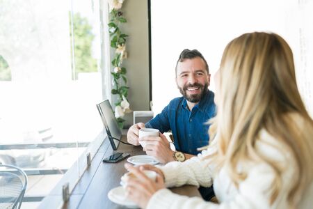 Male and female friends talking while enjoying coffee at desk by window in cafe