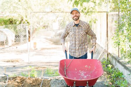 Smiling Hispanic mid adult farmer standing with wheelbarrow at farm on sunny dayの写真素材