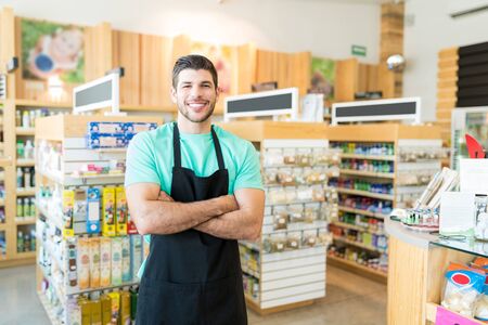 Confident Latin male owner standing with arms crossed in grocery storeの写真素材
