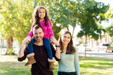 Portrait Of Happy Man Carrying Daughter On Shoulders While Standing By Woman At Parkの写真素材