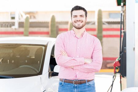 Smiling man in casual standing with arms crossed by car at self-service gas stationの写真素材