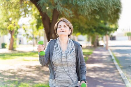 Smiling active elderly Caucasian woman walking while lifting dumbbells in cityの写真素材