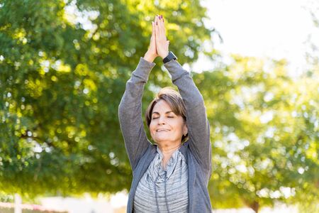 Smiling elderly active woman exercising with arms raised and eyes closed at parkの写真素材