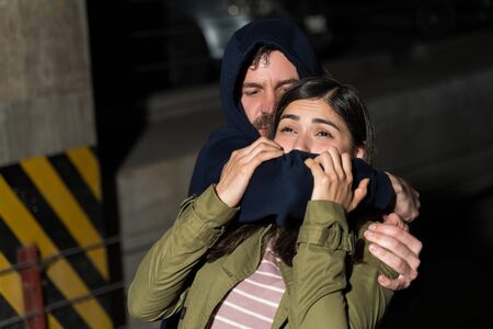 Male thief assaulting female victim while covering her mouth in dark alleyの写真素材