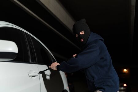Criminal in ski mask opening car door with crowbar while looking away in dark parking lot