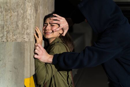 Male criminal using force to coerce female victim while pressing her against wall in parking lotの写真素材
