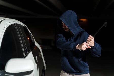 Male criminal breaking car window with crowbar in dark parking lotの写真素材