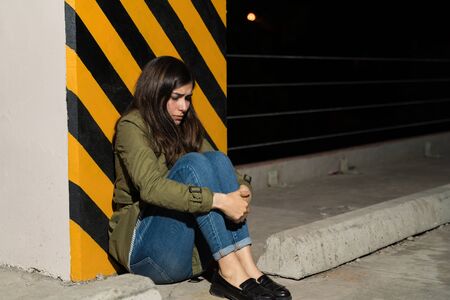 Full length of sad woman hugging knees while sitting against striped column in parking lot at nightの写真素材