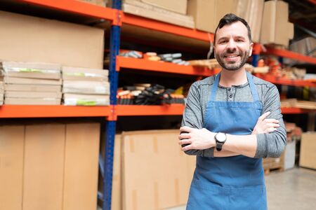 Smiling handsome salesman standing with arms crossed while making eye contact against rack at hardware storeの写真素材
