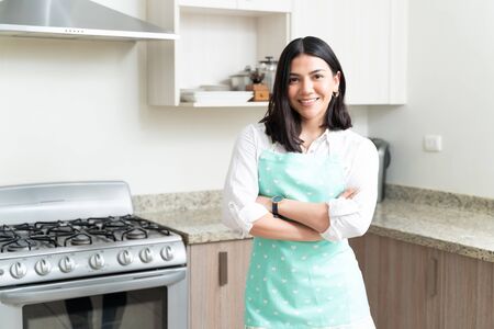 Smiling attractive housewife with arms crossed while standing against stove in kitchenの写真素材