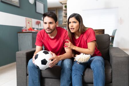 Worried boyfriend and girlfriend watching soccer match in living room at homeの写真素材