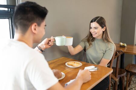 Good looking young couple making a toast with coffee cups in a restaurantの写真素材