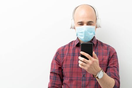 Caucasian man wearing headphones and a surgical mask looking at his smartphone in a studio