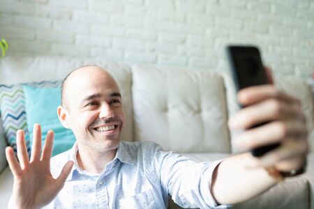 Man in his 30s waving hello with his hand while doing a video call with his smartphone at home and...