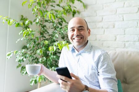 Portrait of a happy Caucasian man reading the news on his phone and enjoying a cup of coffee at home
