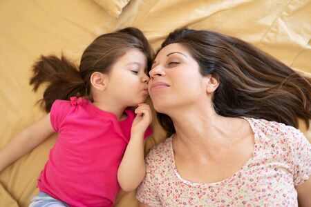 Top view of a pretty little girl kissing her mother on the cheek while they both relax in a bedの写真素材