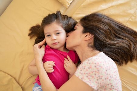 Portrait of a beautiful Caucasian woman in pajamas getting a kiss on the cheek from her mother in bedの写真素材