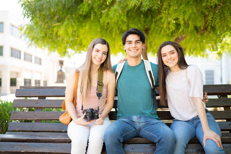 Smiling Latin young man sitting with women on bench in park during weekend tripの写真素材