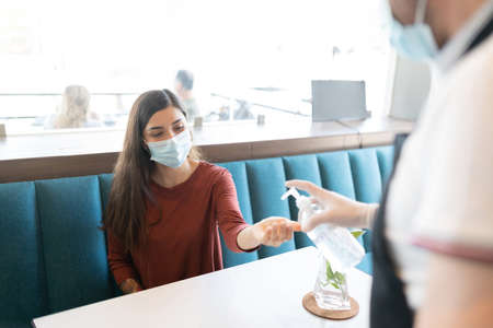 Worker pouring sanitizer on hand of customer sitting at table in cafeの写真素材