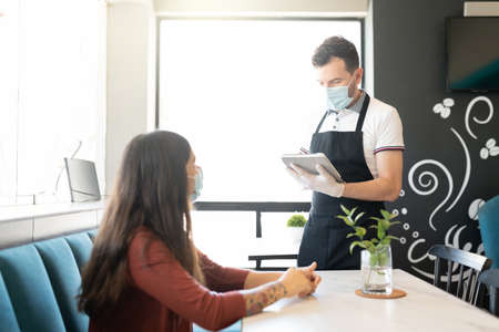 Young woman giving order to waiter wearing mask and gloves during coronavirus outbreak in restaurantの写真素材