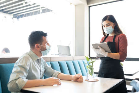 Mid adult man giving order to waitress wearing mask and gloves during coronavirus outbreak in coffee...